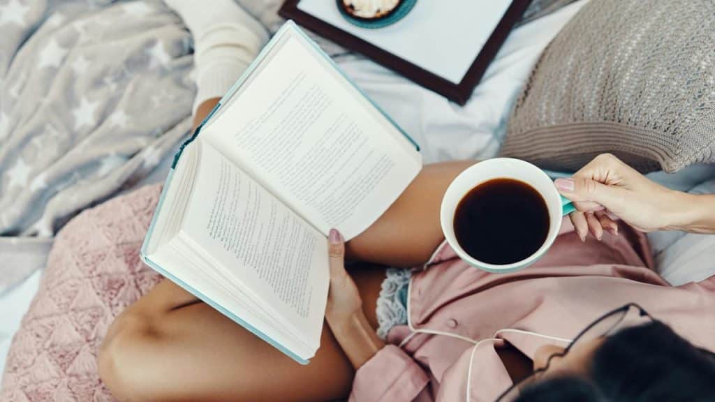 A person reading a book while holding a cup of coffee in bed.
