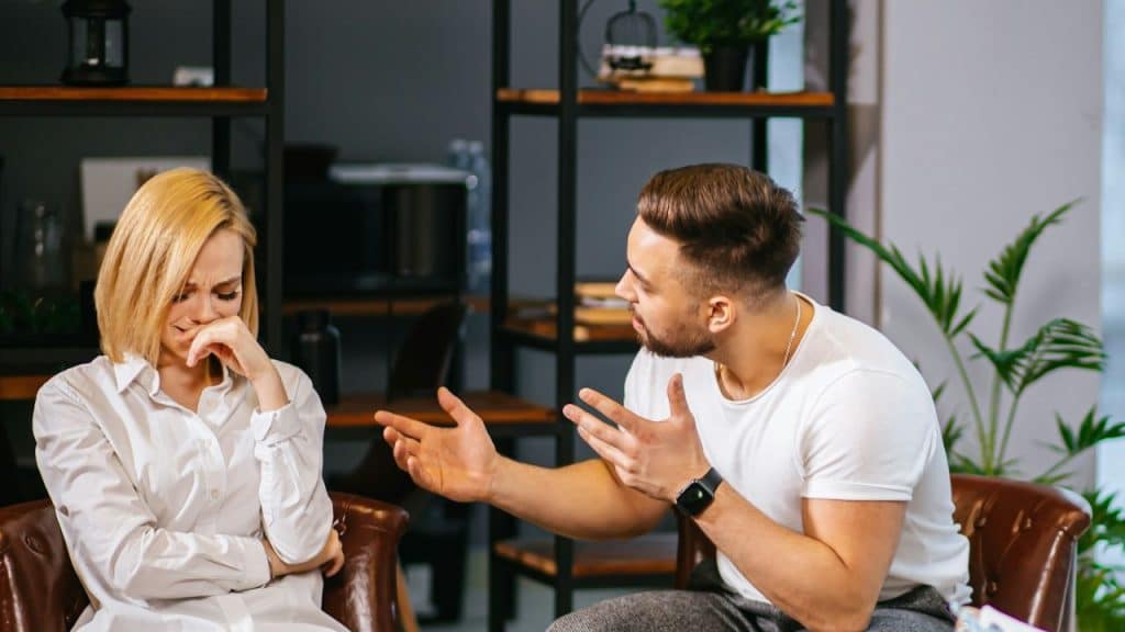 A crying blonde woman sits while a man in a white shirt argues, gesturing with his hands.