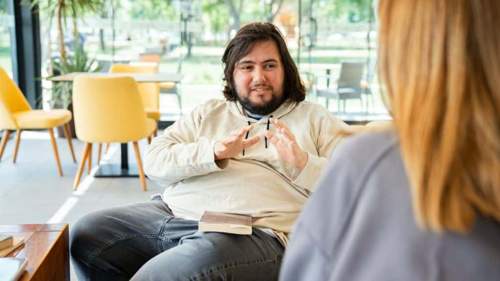 A smiling, long-haired man in a hoodie gestures with his hands while talking to someone indoors.
