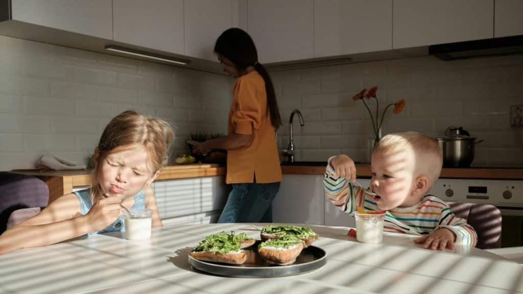 Two children eating at a kitchen table while a woman works in the background.