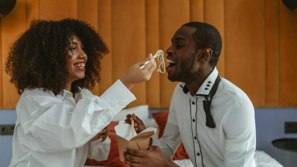 A woman playfully feeding noodles to a man while sitting together indoors.