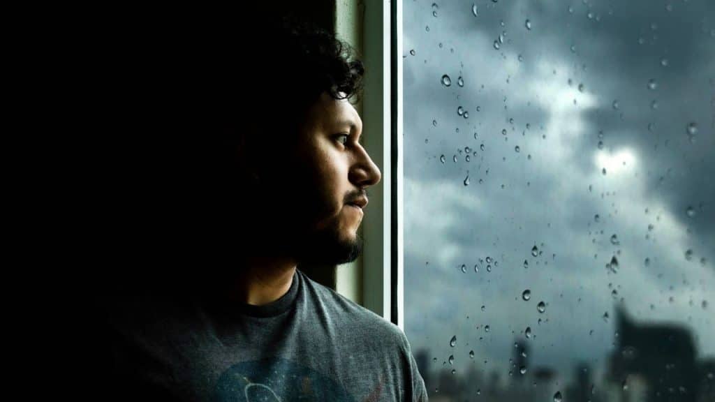 Man standing in the shadows, looking out a window on a rainy day.