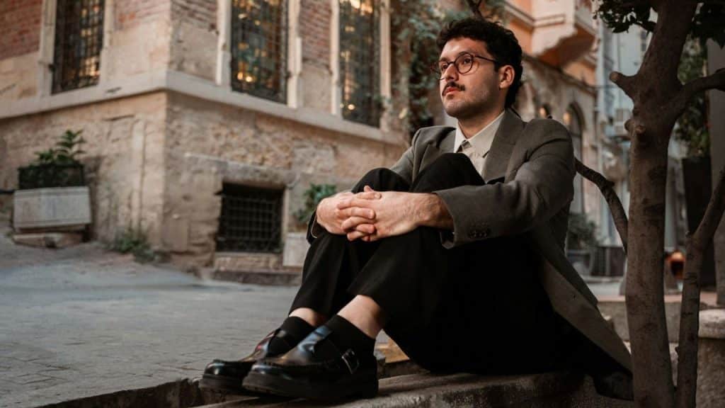 A serious man in a suit jacket and glasses sits alone on a stone step outdoors.
