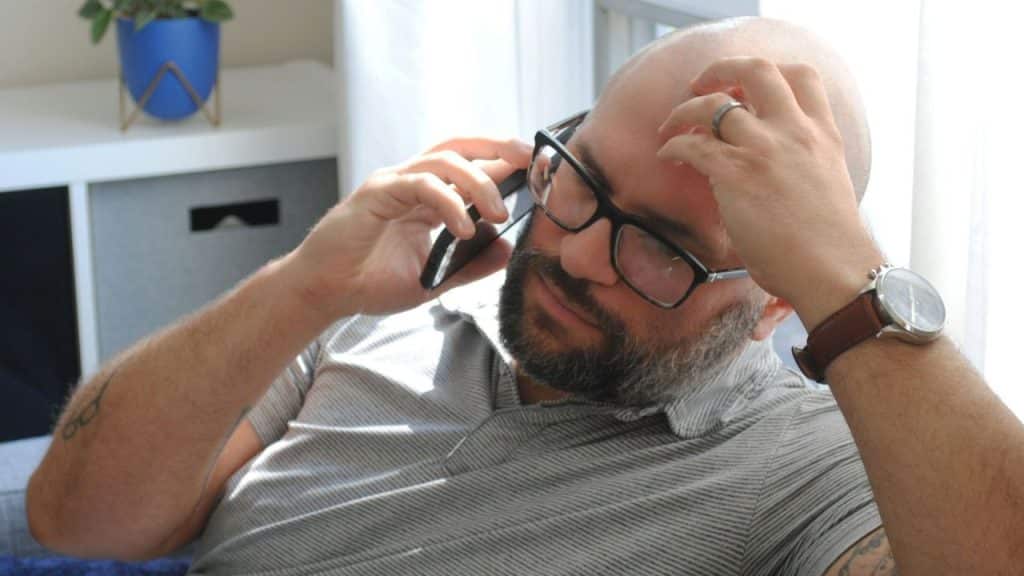 Bald, bearded man wearing glasses talking on a phone indoors, hand on his forehead.