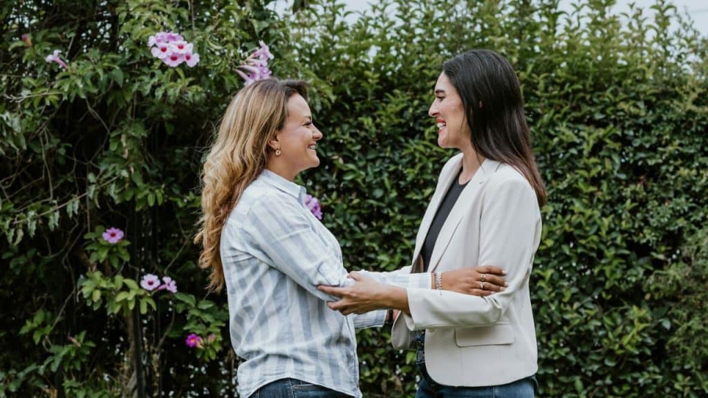 Two women smiling at each other and holding forearms in a garden with green hedges and pink flowers.
