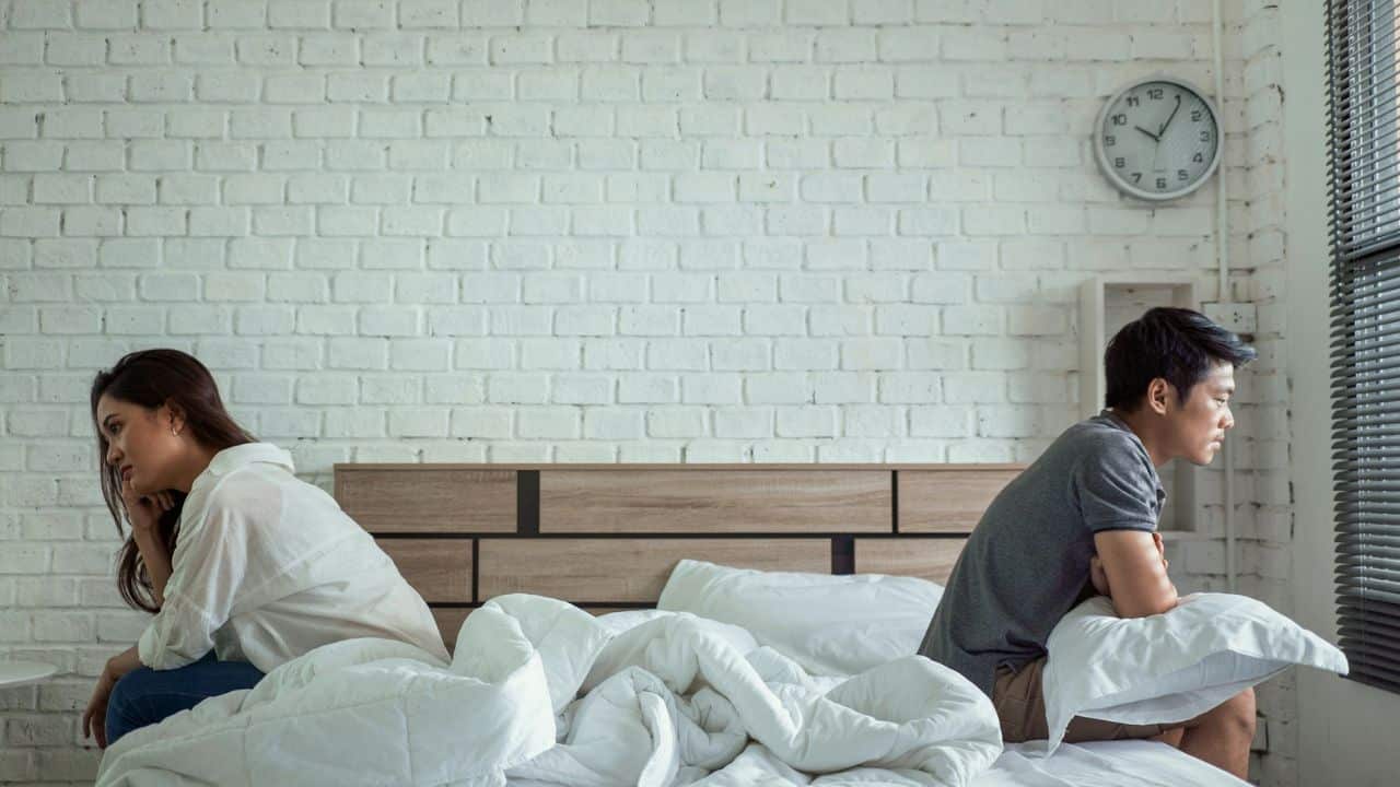 A couple sits separately on a bed with their backs turned to each other.