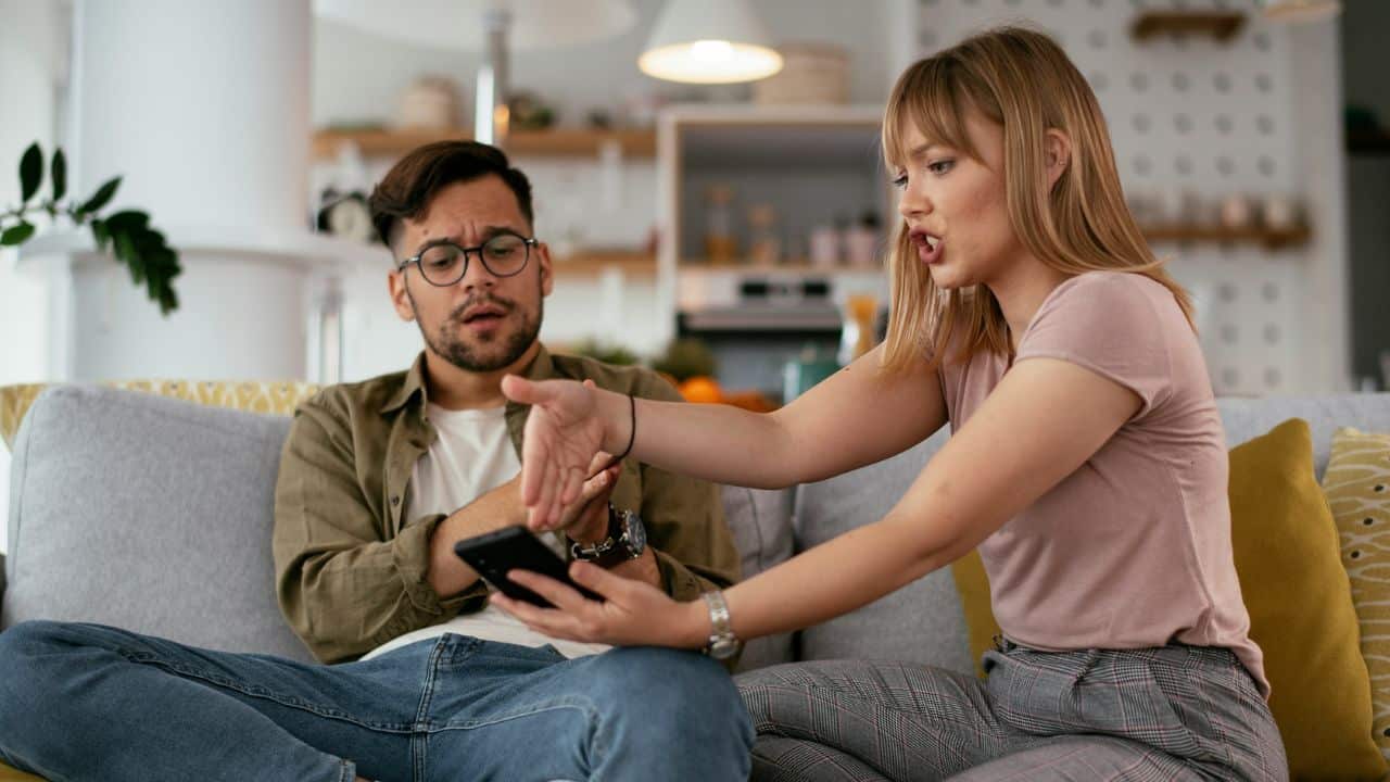 Woman arguing and pointing at a phone held by a concerned man on a sofa.