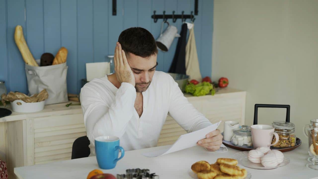 Man in a white shirt looking at a paper, holding his head, with food on the table.