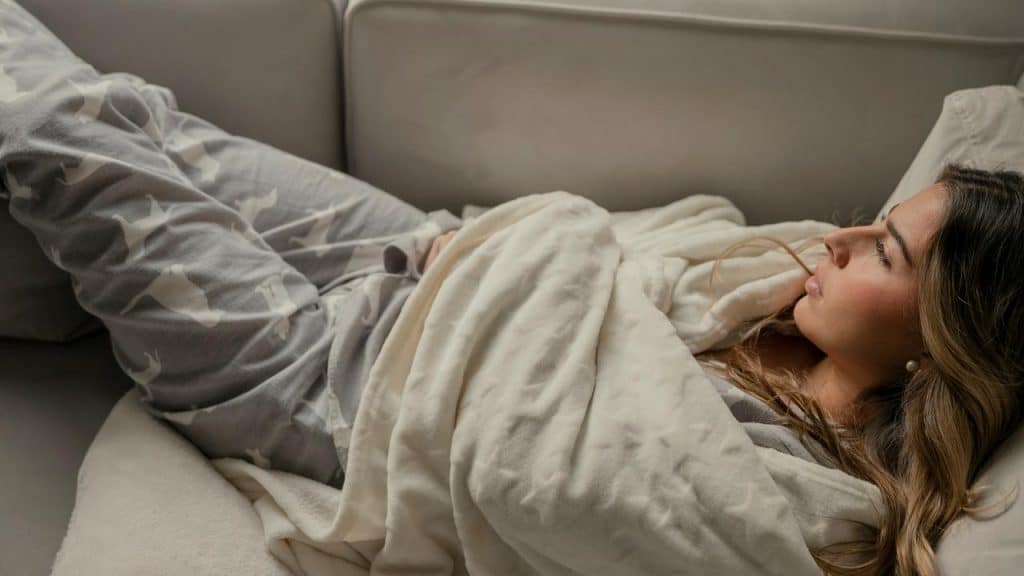 A person wearing gray pajamas lies on a couch under a soft cream blanket.
