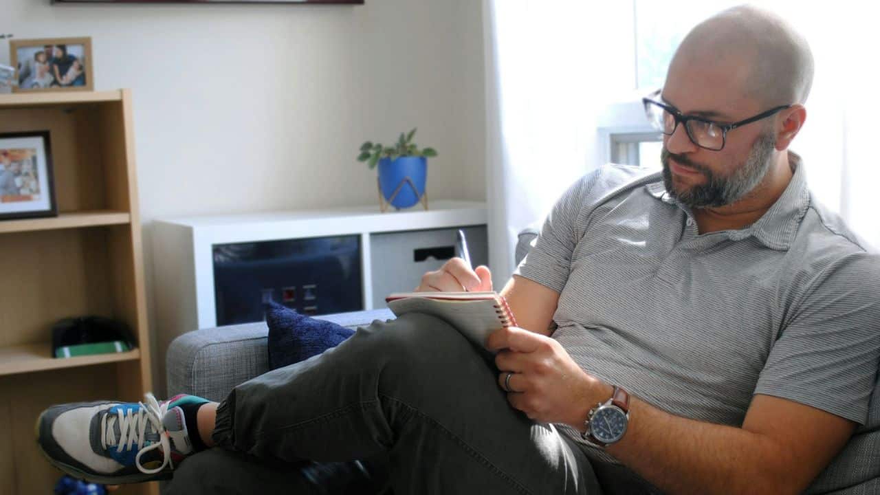 Bald man with a beard and glasses writing in a spiral notebook in a living room.