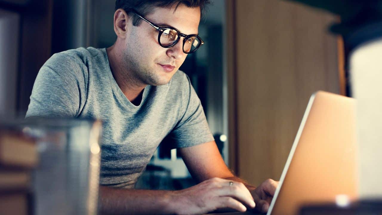 Young man in glasses and a grey shirt working intently on a laptop indoors.