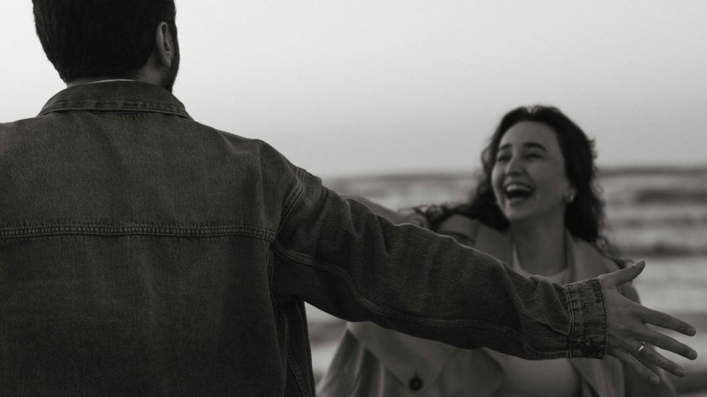 A man greeting a woman with open arms while she laughs.