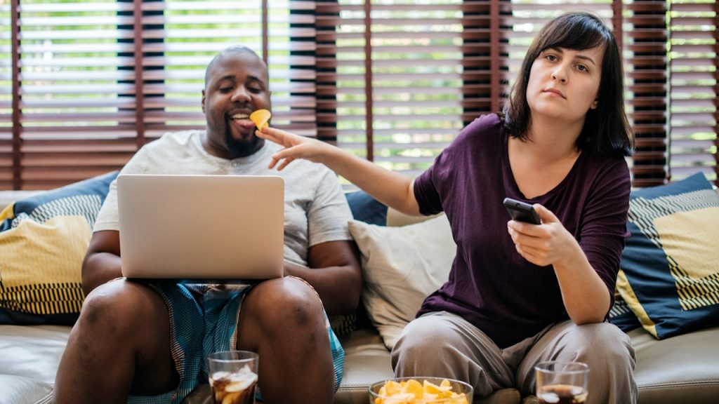 A woman feeding a chip to a man working on a laptop while she uses the phone with an annoyed expression.