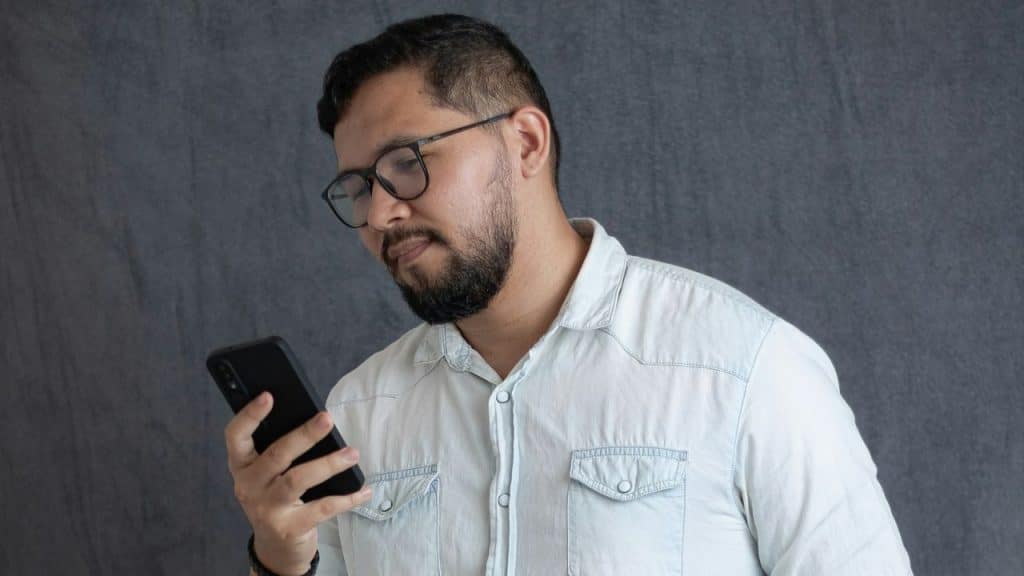 Bearded man with glasses and a light denim shirt looking at his smartphone.