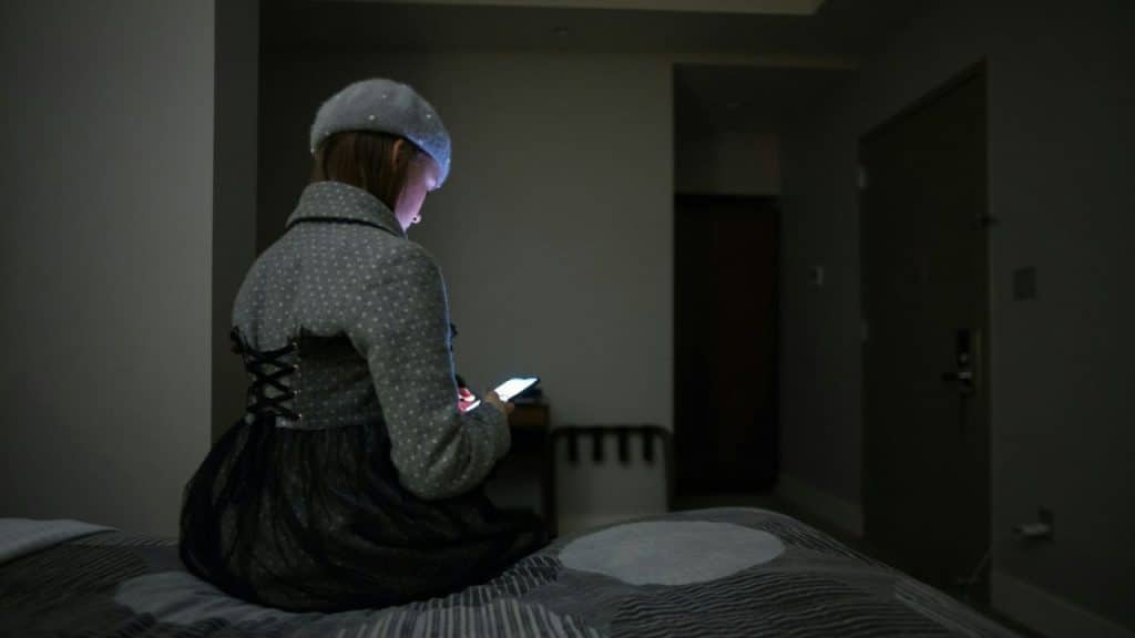 Woman in a jacket and hat sitting on a bed in a dark room looking at a glowing phone.