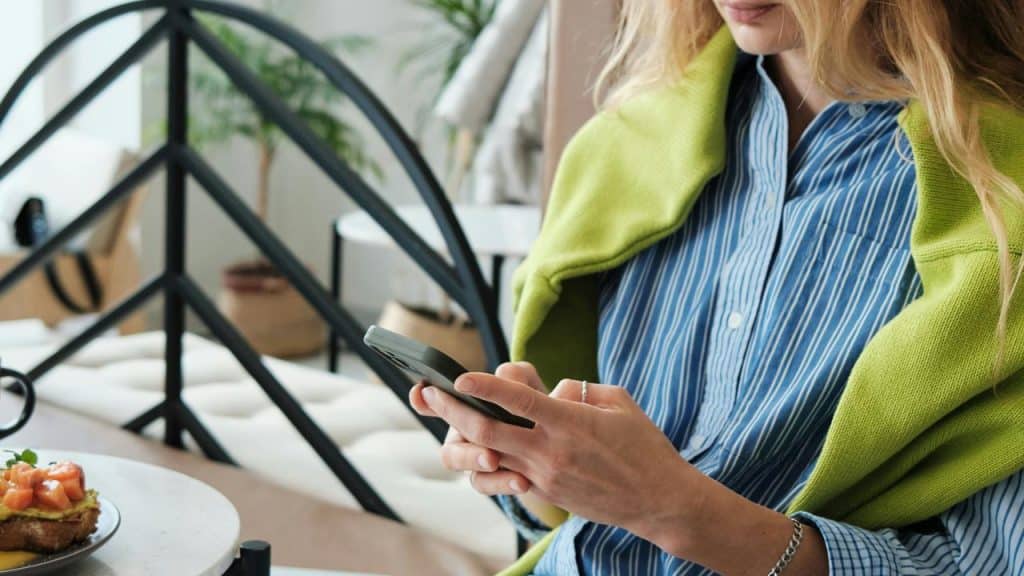 A woman in a striped shirt and green sweater uses her phone at a table with food.