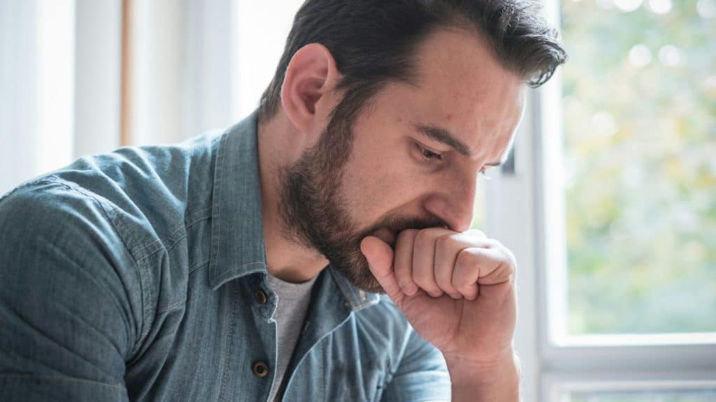 Pensive bearded man in a denim shirt holding his fist near his mouth indoors.