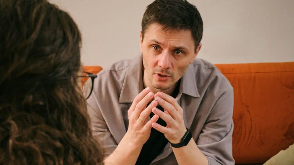 A man in a gray shirt talking earnestly with his hands clasped to a person across from him.