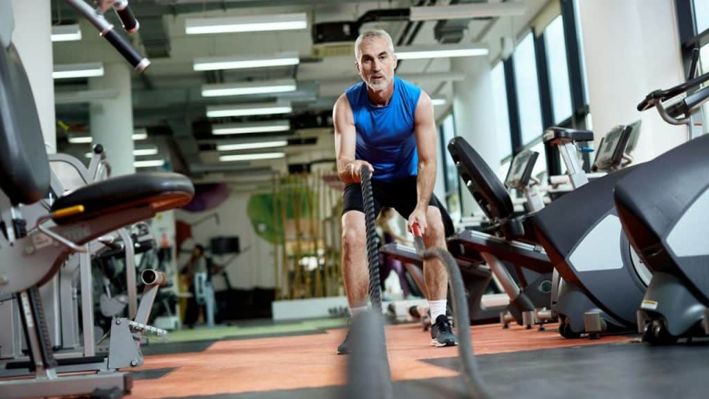 Older man in a blue tank top exercising with battle ropes in a large gym.