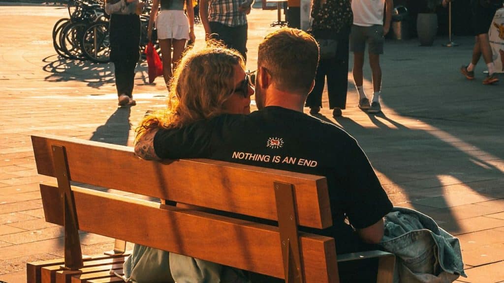 Couple sitting on a wooden bench, embracing, with people walking in the background.