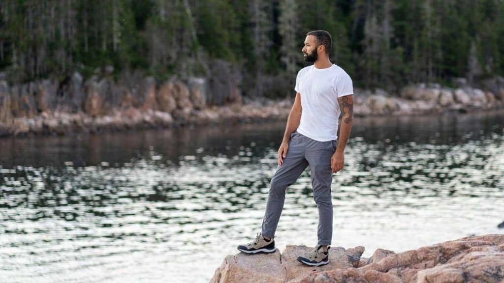 Man in a white t-shirt and gray pants standing on a rock by a body of water, looking away.