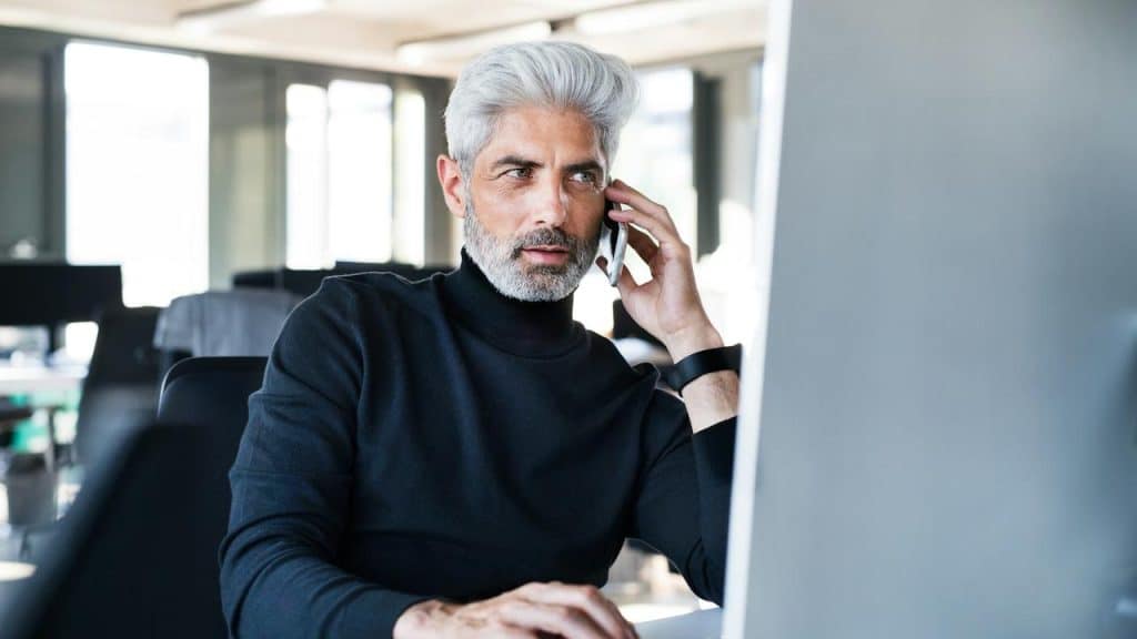A man with gray hair and beard talking on the phone while working at a computer.