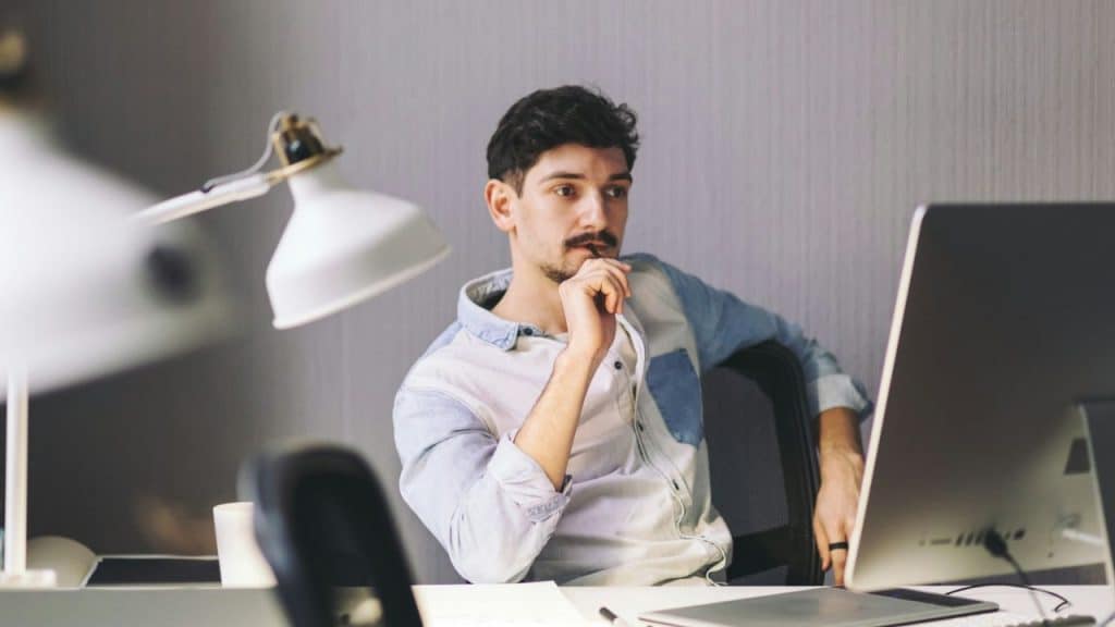 Young man with a mustache sitting at a desk, looking thoughtfully at a computer monitor.