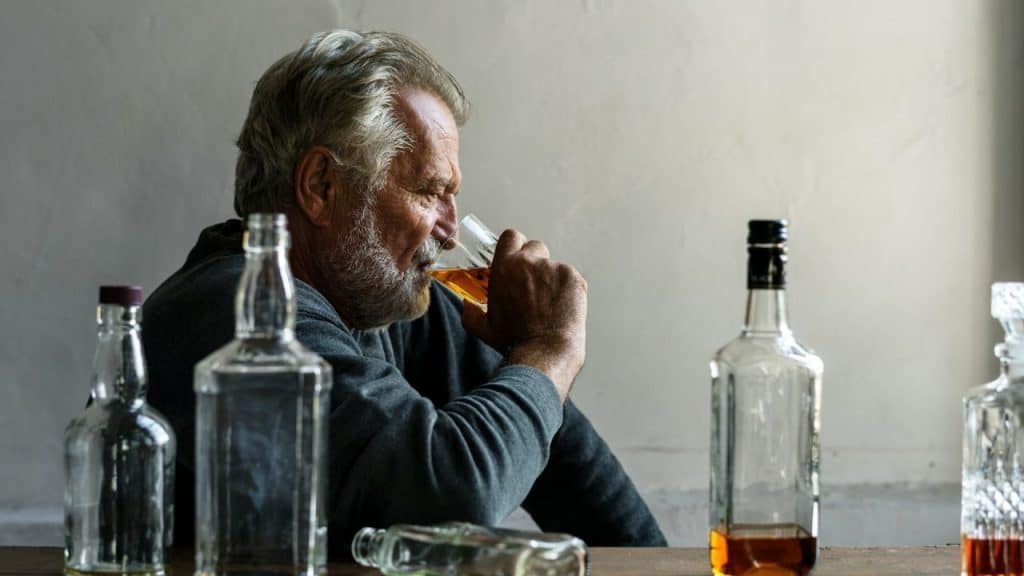 Older bearded man sitting at a table surrounded by liquor bottles, drinking from a glass.