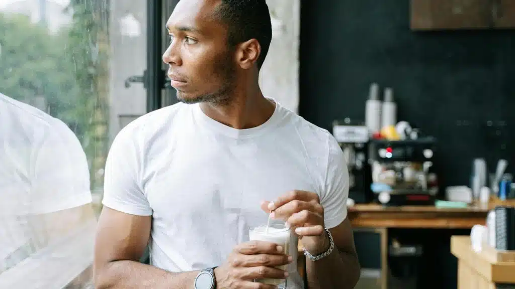 A man in a white t-shirt holding a drink while looking out the window.