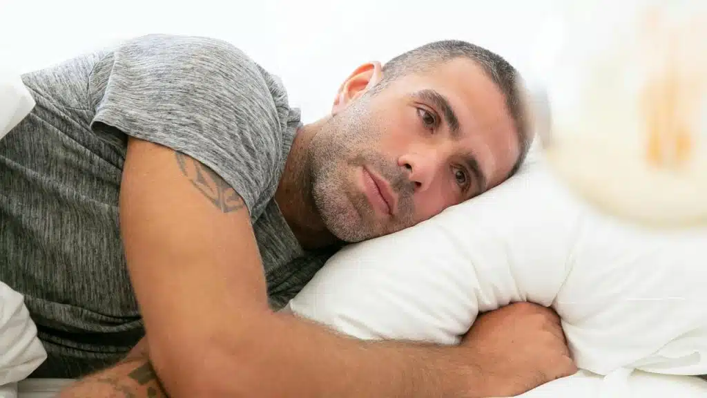 A man lying on a bed with his head on a pillow, looking thoughtful.