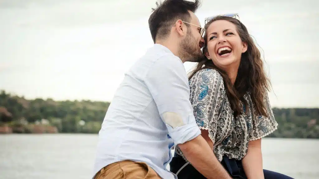 A man is about to kiss a laughing woman on the cheek while they sit in front of a lake.