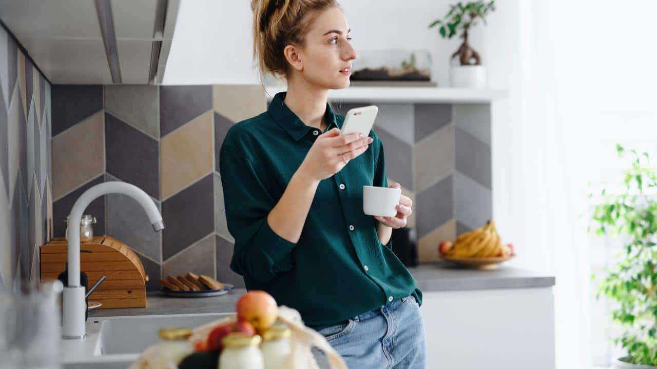 A woman in a kitchen holding a phone and a cup while looking to the side.