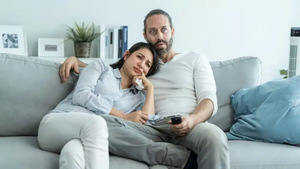 A couple sits on a sofa watching television with concerned, uneasy expressions.