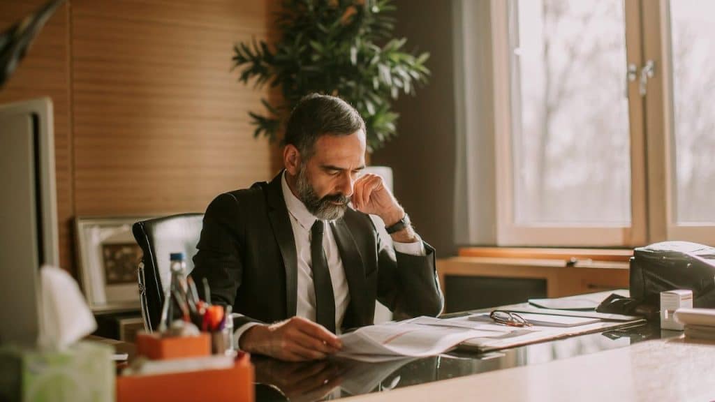A man in a suit with a beard sits at a desk, reviewing papers in a sunlit office.