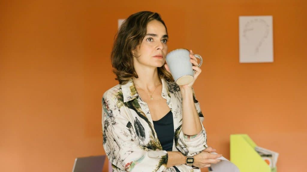 A woman with short brown hair holds a mug and looks away from the camera against an orange wall.