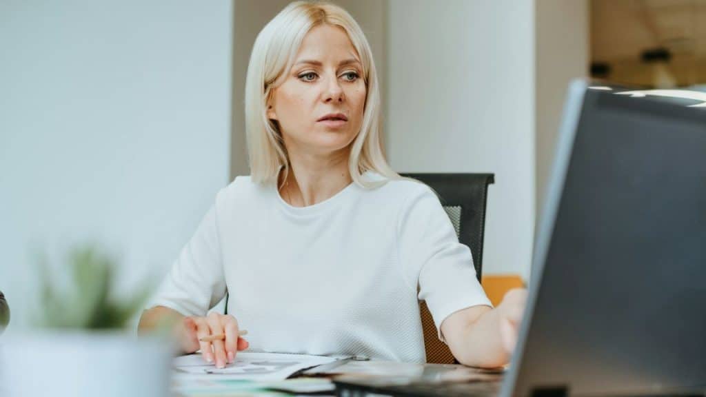 A blonde woman in a white shirt intently looks at a laptop screen while sitting at a desk.