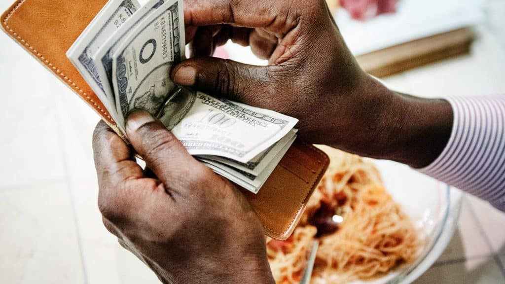 A person holds a wallet with several US dollar bills near a plate of food.