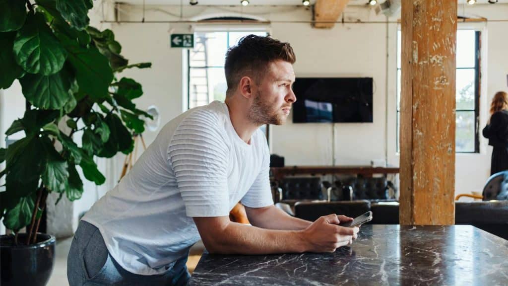 A man in a white shirt leans on a table while holding a phone, gazing thoughtfully.