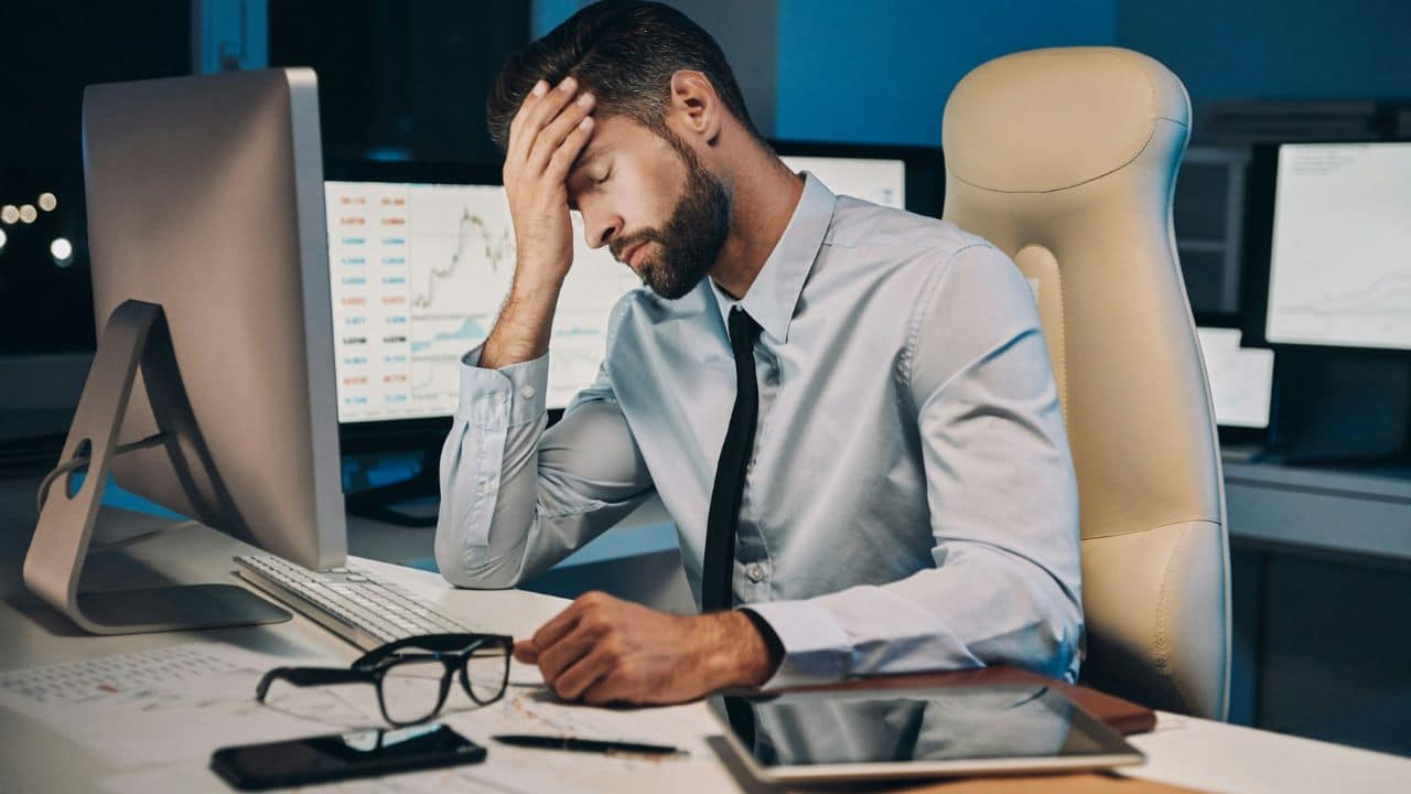 A man in a shirt and tie, exhausted at his computer, holding his forehead.
