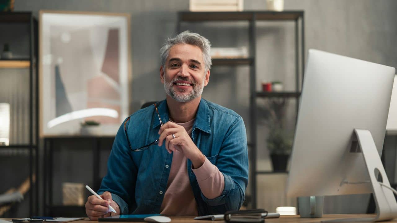 A smiling man with gray hair is sitting at a desk with a computer and tablet.