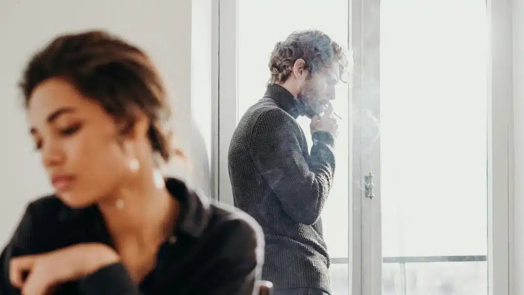 A man is standing by a bright window, smoking a cigarette, while a woman sits in the foreground, looking down.