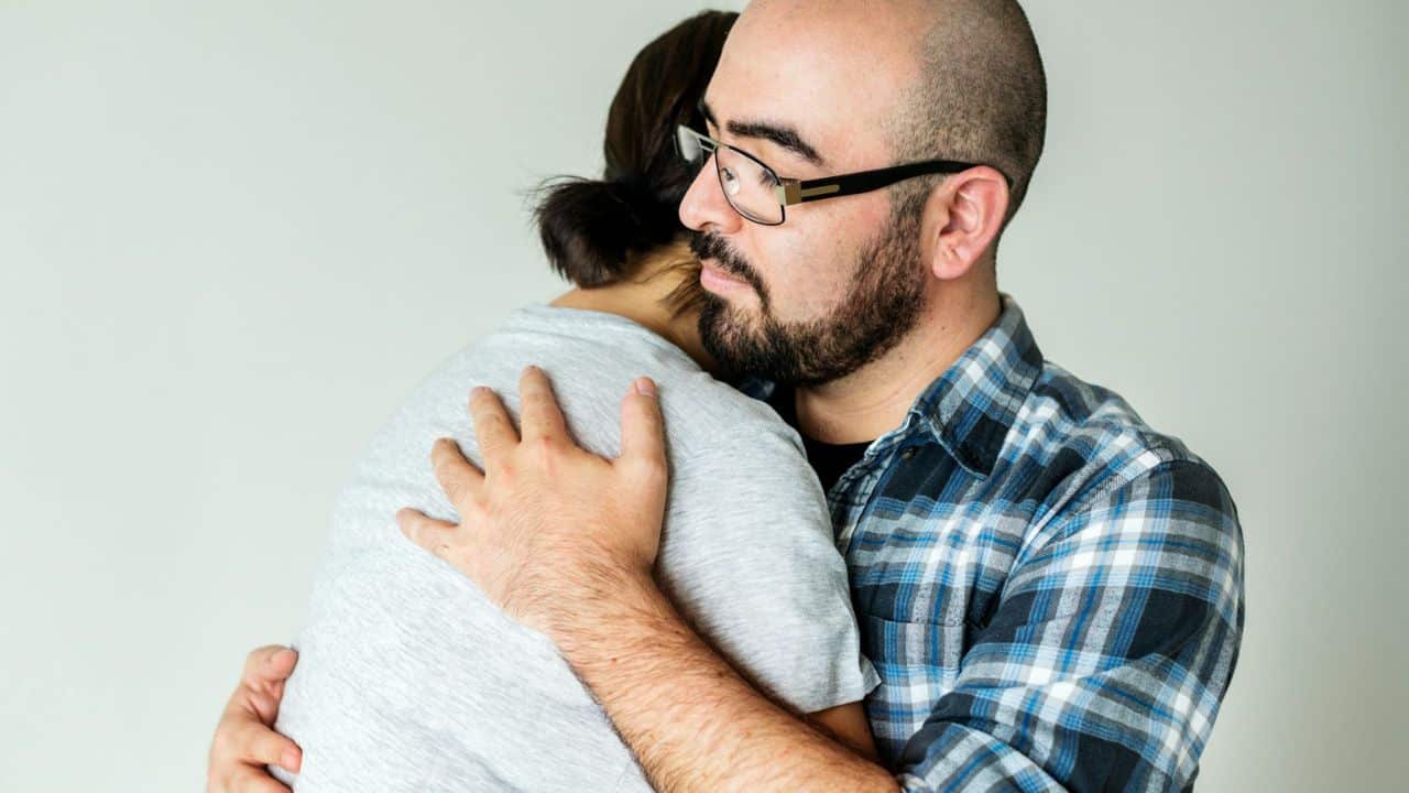 A bearded man wearing glasses and a plaid shirt is hugging a woman in a gray shirt.