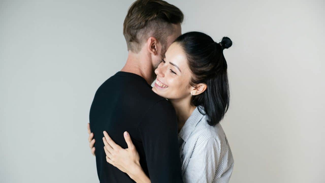 A woman with a happy smile is hugging a man against a plain light background.