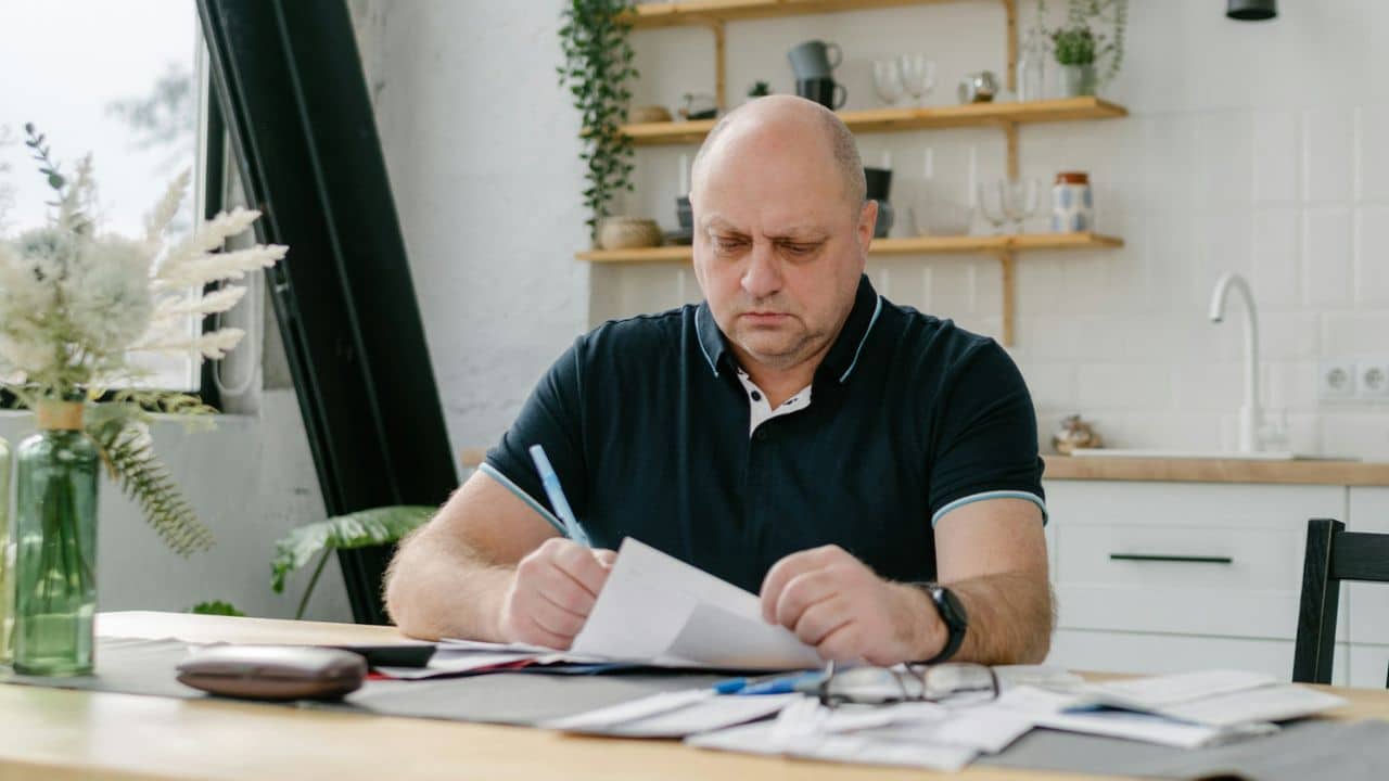 A bald man in a dark polo shirt writing on papers at a kitchen table.