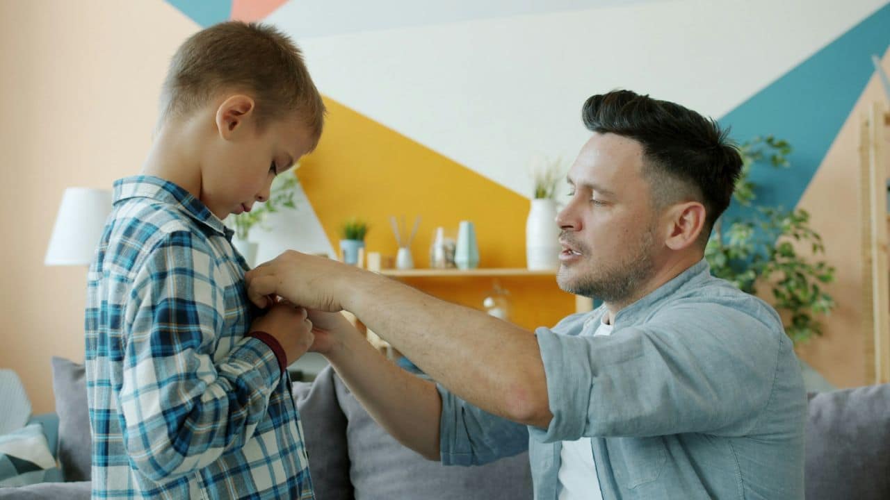 A father helps his young son button up his plaid shirt indoors.