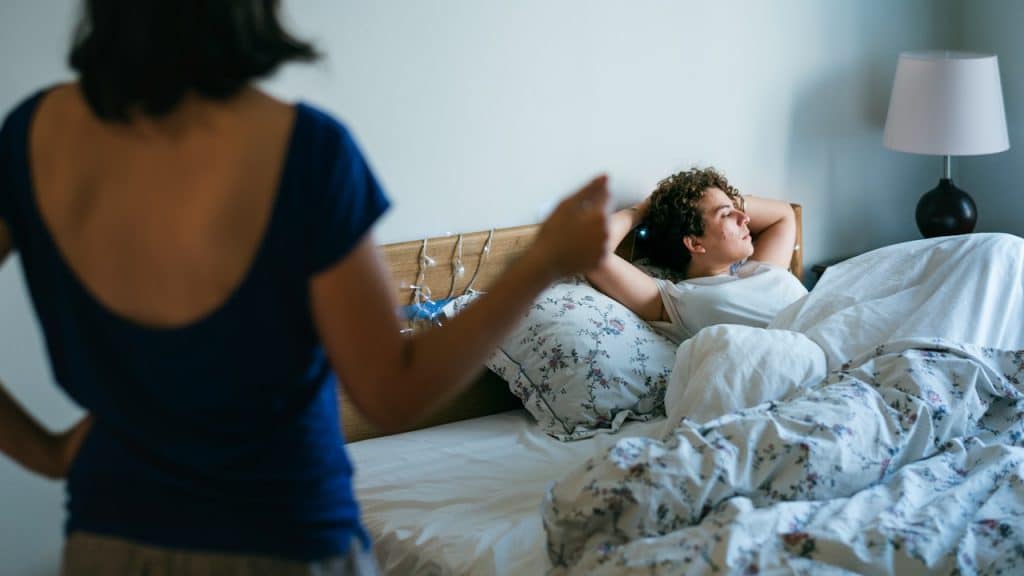 A person lying in bed looking at another person standing by the bed.