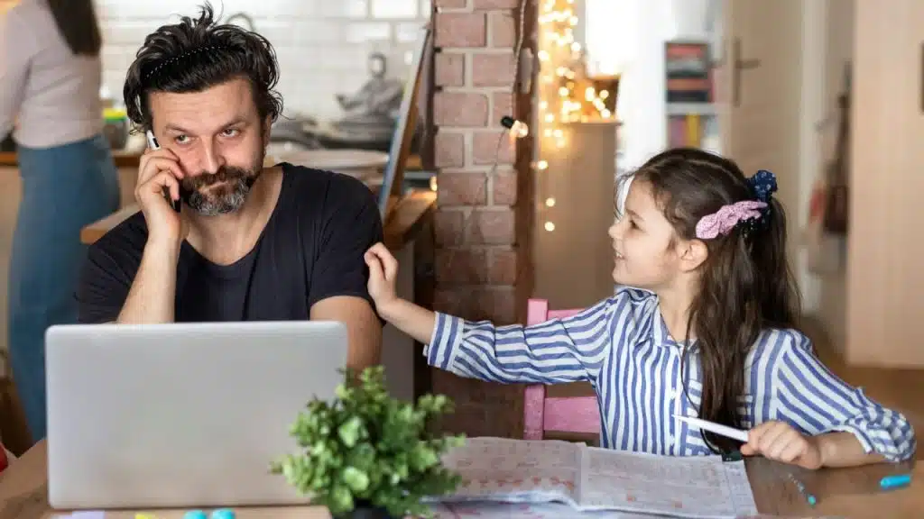A man on a phone call works on a laptop while a girl reaches out to him.