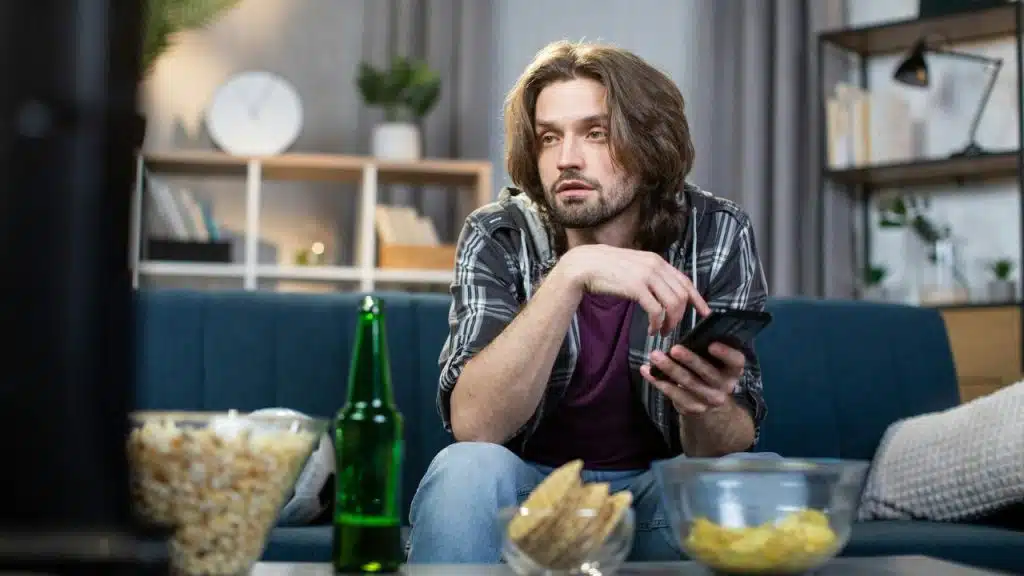 A long-haired man sits on a couch, holding a phone with snacks and beer nearby.