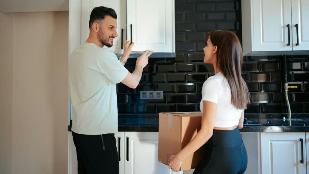 A man smiles while pointing at cabinets as a woman holds a box in a kitchen.