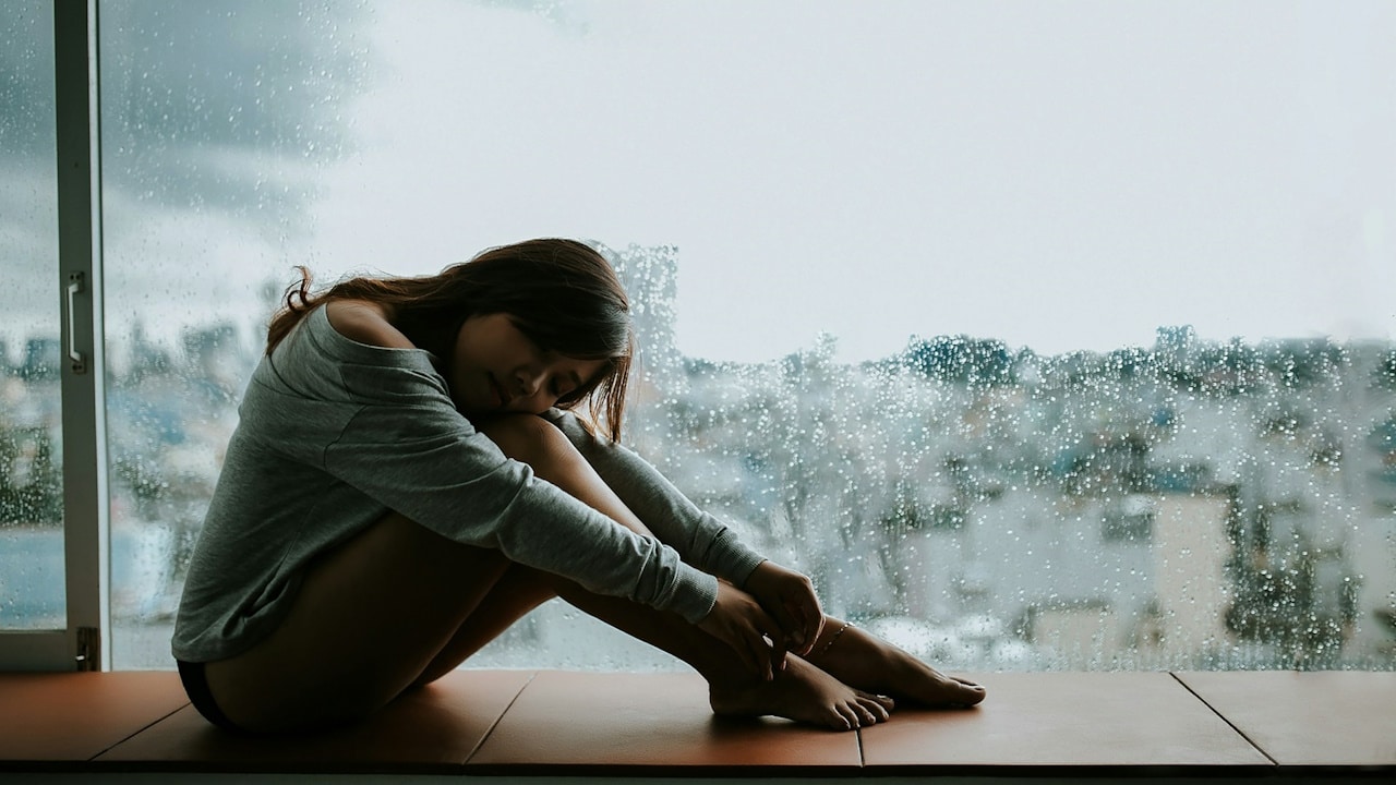 A sad woman wearing a gray shirt is sitting on a window.